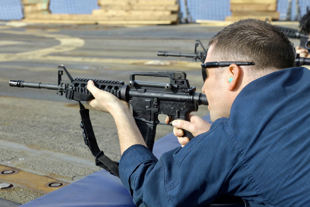 A person in a blue uniform is in a prone or supported shooting position aiming an M4/AR-15 style carbine rifle. The setting appears to be an outdoor range on what looks like a naval vessel or industrial deck. Additional rifles and at least one other shooter are visible in the background.