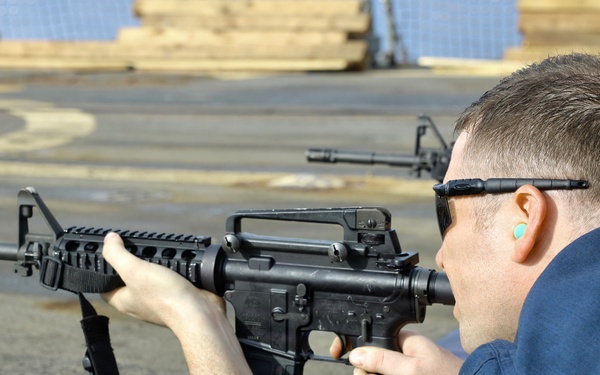 A person in a blue uniform is in a prone or supported shooting position aiming an M4/AR-15 style carbine rifle. The setting appears to be an outdoor range on what looks like a naval vessel or industrial deck. Additional rifles and at least one other shooter are visible in the background.