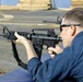 A person in a blue uniform is in a prone or supported shooting position aiming an M4/AR-15 style carbine rifle. The setting appears to be an outdoor range on what looks like a naval vessel or industrial deck. Additional rifles and at least one other shooter are visible in the background.
