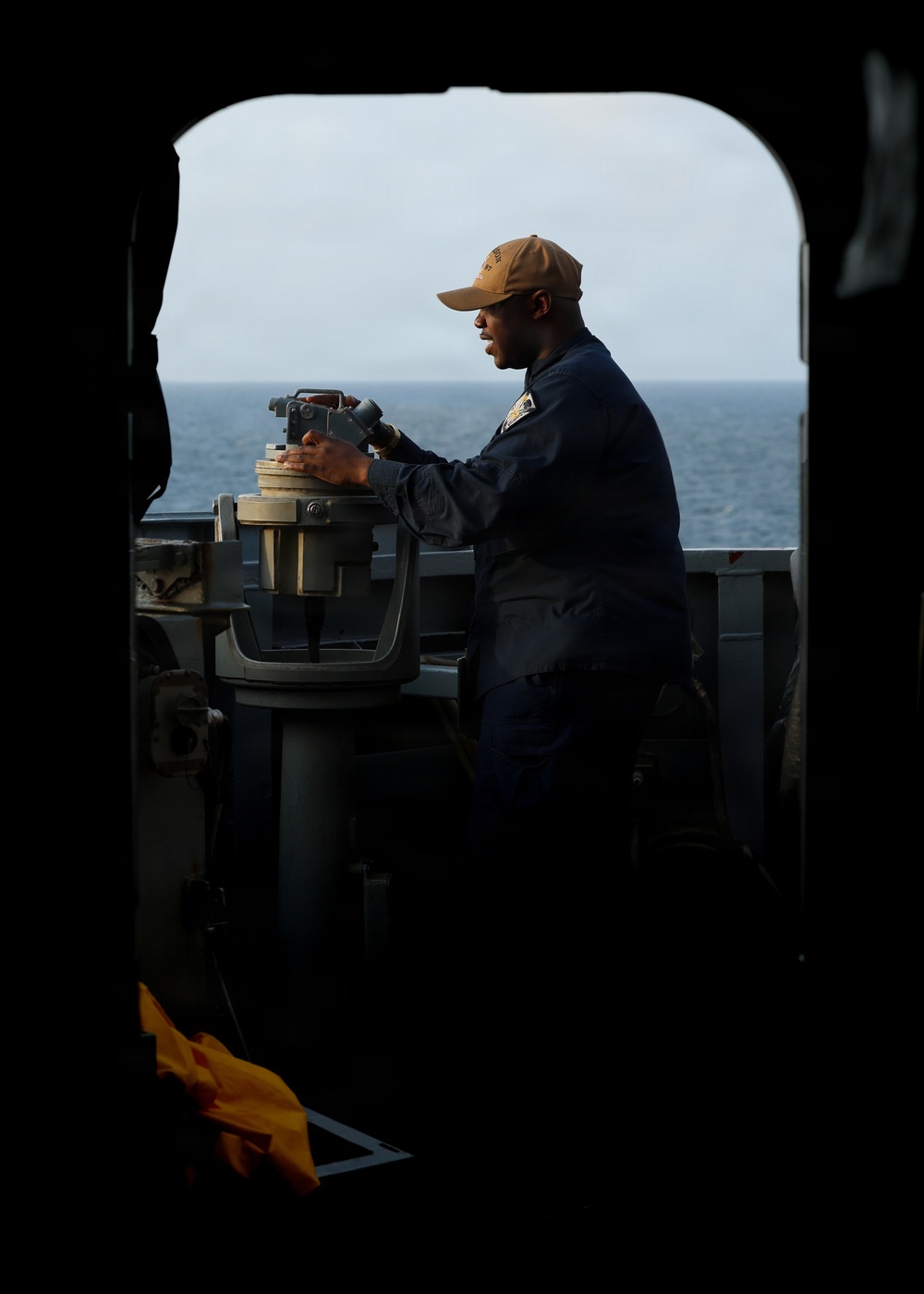 A naval service member in dark blue coveralls and a tan cap is operating a gyrocompass or bearing circle mounted on a pedestal on the bridge wing of a naval vessel. The image is framed through a hatch or doorway opening, creating a dramatic silhouette effect with the open ocean visible in the background. The photograph appears to be taken aboard a U.S. Navy ship at sea.
