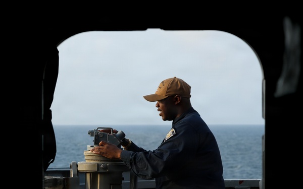 A naval service member in dark blue coveralls and a tan cap is operating a gyrocompass or bearing circle mounted on a pedestal on the bridge wing of a naval vessel. The image is framed through a hatch or doorway opening, creating a dramatic silhouette effect with the open ocean visible in the background. The photograph appears to be taken aboard a U.S. Navy ship at sea.