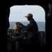 A naval service member in dark blue coveralls and a tan cap is operating a gyrocompass or bearing circle mounted on a pedestal on the bridge wing of a naval vessel. The image is framed through a hatch or doorway opening, creating a dramatic silhouette effect with the open ocean visible in the background. The photograph appears to be taken aboard a U.S. Navy ship at sea.