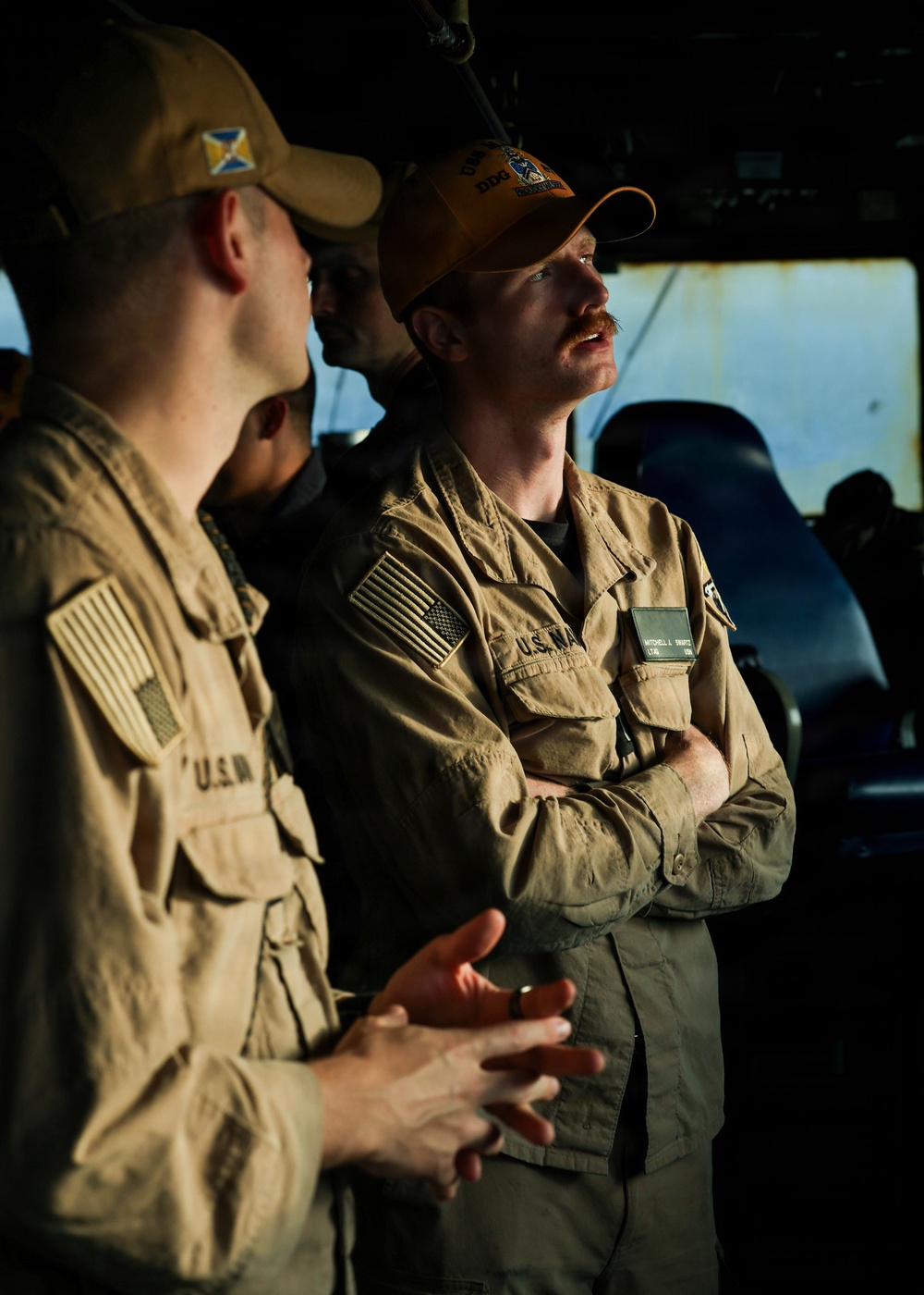 Two U.S. Navy personnel in tan/khaki uniforms are engaged in conversation aboard what appears to be a ship's bridge or interior compartment. The individual on the right wears an orange ball cap with 'DDG' visible and has a mustache, with arms crossed. Both wear U.S. Navy insignia and American flag patches on their sleeves.