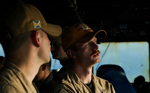 Two U.S. Navy personnel in tan/khaki uniforms are engaged in conversation aboard what appears to be a ship's bridge or interior compartment. The individual on the right wears an orange ball cap with 'DDG' visible and has a mustache, with arms crossed. Both wear U.S. Navy insignia and American flag patches on their sleeves.