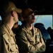 Two U.S. Navy personnel in tan/khaki uniforms are engaged in conversation aboard what appears to be a ship's bridge or interior compartment. The individual on the right wears an orange ball cap with 'DDG' visible and has a mustache, with arms crossed. Both wear U.S. Navy insignia and American flag patches on their sleeves.