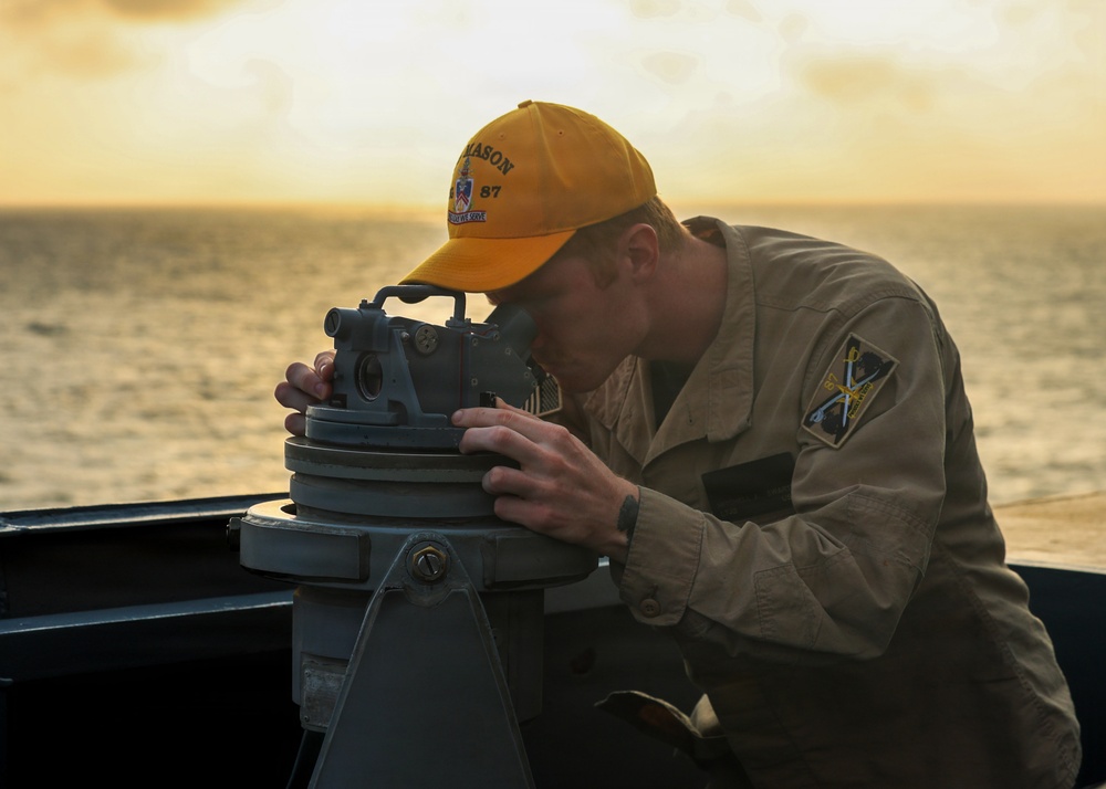 A naval service member is using a gyrocompass or pelorus (bearing circle) mounted on the deck of a ship, likely taking a navigational bearing or celestial observation. The scene is set during golden hour with an ocean horizon visible in the background. The individual is wearing a tan/khaki uniform with a yellow cap bearing the text 'MASON 87'.