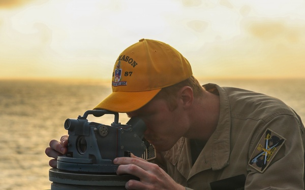 A naval service member is using a gyrocompass or pelorus (bearing circle) mounted on the deck of a ship, likely taking a navigational bearing or celestial observation. The scene is set during golden hour with an ocean horizon visible in the background. The individual is wearing a tan/khaki uniform with a yellow cap bearing the text 'MASON 87'.