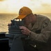A naval service member is using a gyrocompass or pelorus (bearing circle) mounted on the deck of a ship, likely taking a navigational bearing or celestial observation. The scene is set during golden hour with an ocean horizon visible in the background. The individual is wearing a tan/khaki uniform with a yellow cap bearing the text 'MASON 87'.