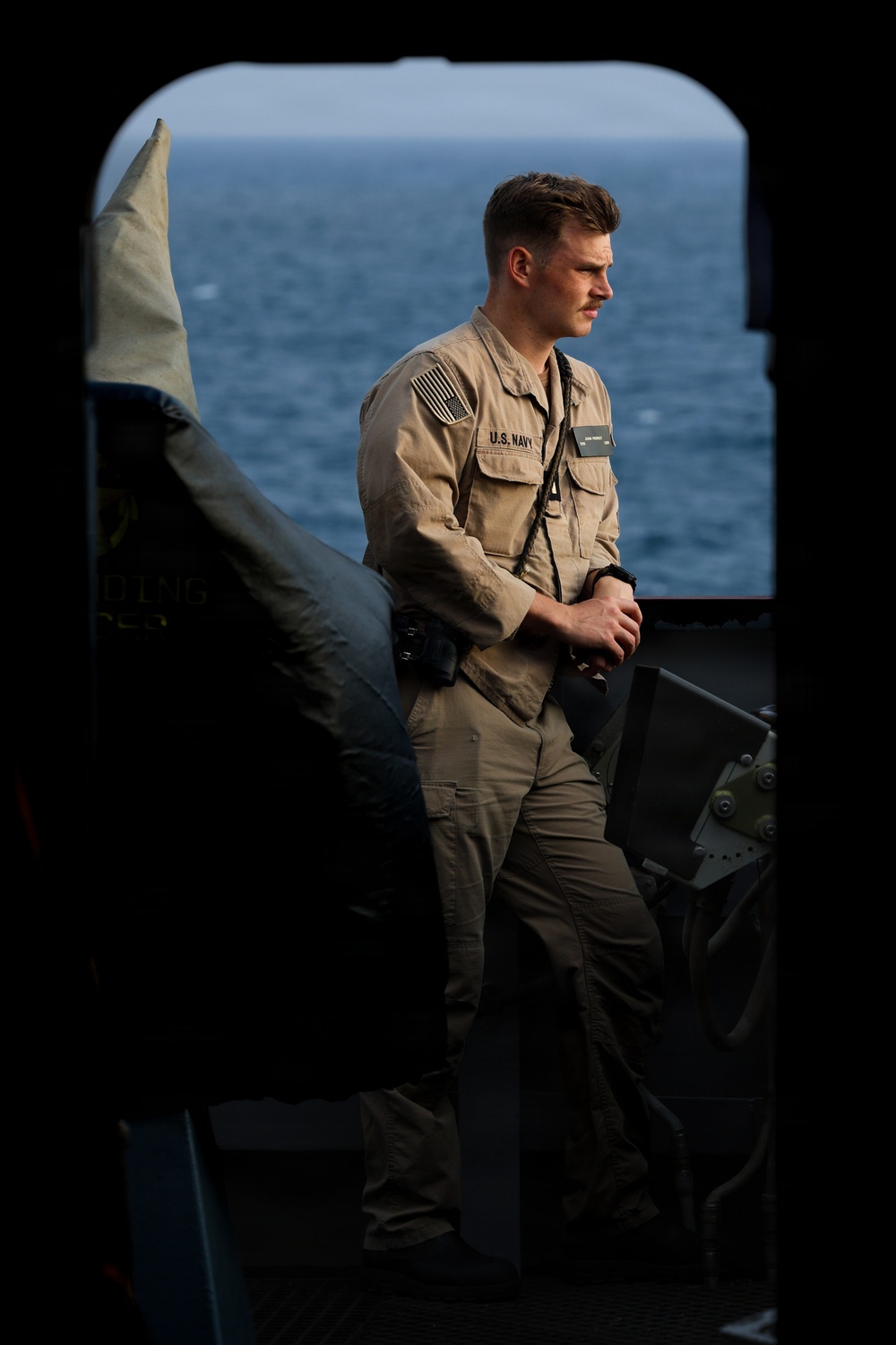 A U.S. Navy sailor in tan/khaki working uniform stands on what appears to be the deck or interior threshold of a naval vessel, with open ocean visible in the background. The sailor is wearing a name tag, U.S. Navy chest tape, an American flag patch on the right shoulder, and a lanyard. The image is framed through a dark archway or hatch opening, creating a dramatic compositional effect.