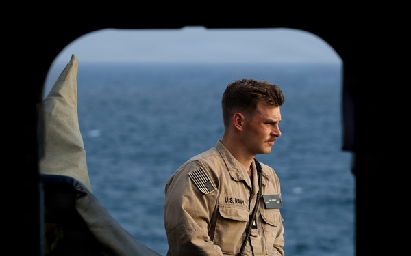 A U.S. Navy sailor in tan/khaki working uniform stands on what appears to be the deck or interior threshold of a naval vessel, with open ocean visible in the background. The sailor is wearing a name tag, U.S. Navy chest tape, an American flag patch on the right shoulder, and a lanyard. The image is framed through a dark archway or hatch opening, creating a dramatic compositional effect.