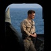 A U.S. Navy sailor in tan/khaki working uniform stands on what appears to be the deck or interior threshold of a naval vessel, with open ocean visible in the background. The sailor is wearing a name tag, U.S. Navy chest tape, an American flag patch on the right shoulder, and a lanyard. The image is framed through a dark archway or hatch opening, creating a dramatic compositional effect.