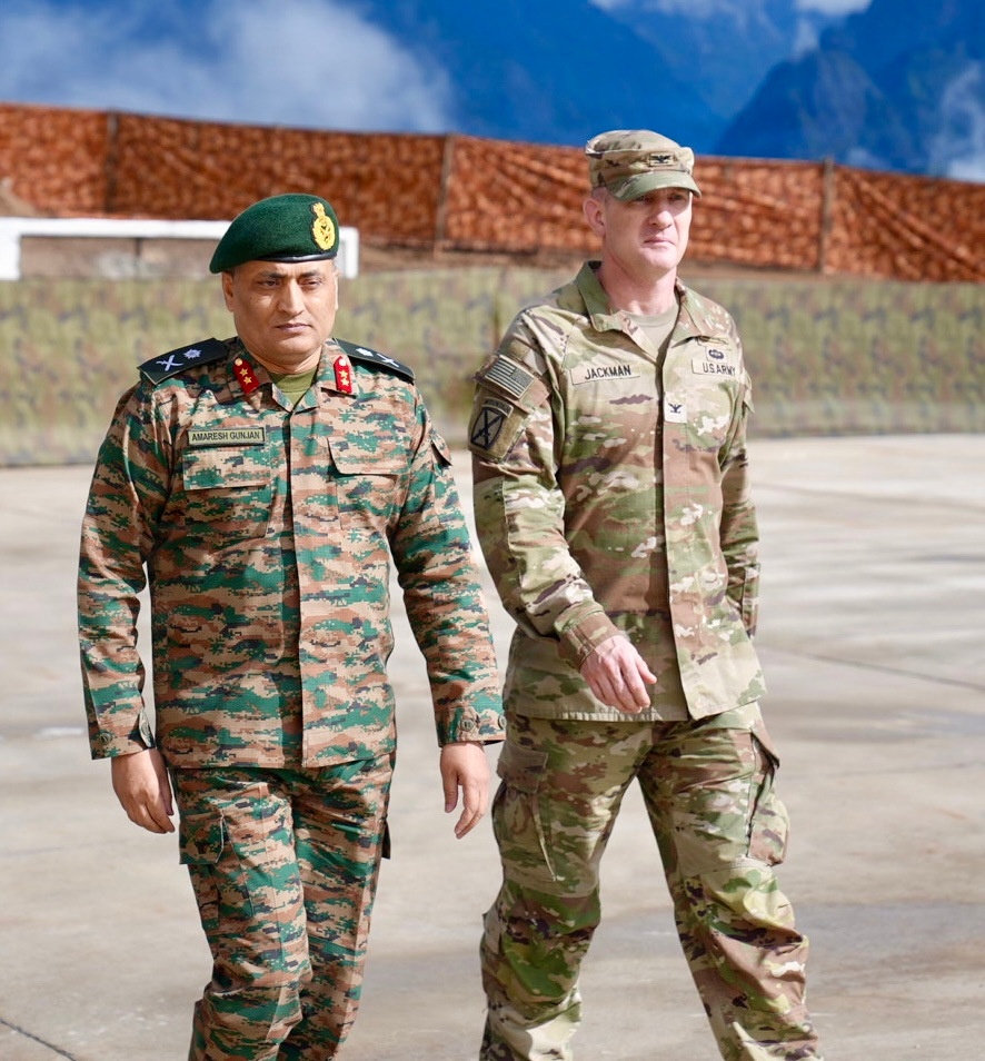 Two military officers — one from the Indian Army and one from the U.S. Army — are walking together on what appears to be a tarmac or parade ground with mountainous terrain visible in the background. The Indian officer wears a green beret and name tape reading 'AMARESH GUNJAN' with lieutenant general rank insignia, while the U.S. officer's name tape reads 'JACKMAN' with 'US ARMY' and a colonel's eagle rank insignia. The setting and uniforms suggest a bilateral military engagement or joint exercise.
