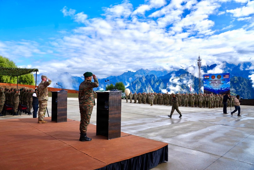 A formal military ceremony for 'Yudh Abhyas XXII,' a joint India-USA military exercise, is taking place on an outdoor stage with mountainous terrain in the background. Two officers — one in Indian Army camouflage with a green beret and one in US Army combat uniform — are saluting at podiums on a raised platform. A large formation of soldiers from both nations stands at attention in the background.