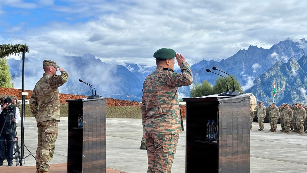Two military officers from different nations are saluting at podiums during what appears to be a formal joint military ceremony or exercise. One officer wears US Army MultiCam (OCP) uniform, while the other wears a distinctive green beret with a different camouflage pattern, suggesting a multinational military event. The ceremony takes place outdoors against a dramatic mountainous backdrop with snow-capped peaks.