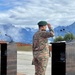 Two military officers from different nations are saluting at podiums during what appears to be a formal joint military ceremony or exercise. One officer wears US Army MultiCam (OCP) uniform, while the other wears a distinctive green beret with a different camouflage pattern, suggesting a multinational military event. The ceremony takes place outdoors against a dramatic mountainous backdrop with snow-capped peaks.