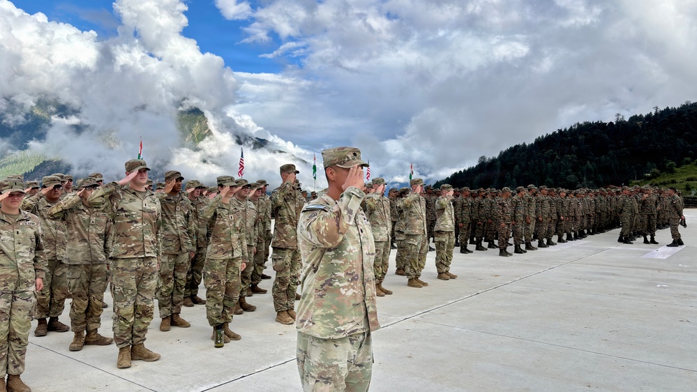 A military parade or ceremony is taking place outdoors at a mountainous location, featuring two distinct groups of soldiers in formation facing each other. Soldiers on the left wear US Army OCP (Operational Camouflage Pattern) uniforms, while soldiers on the right appear to wear a different camouflage pattern, suggesting a joint multinational military exercise or ceremony. A central figure in OCP uniform is rendering a salute, and multiple national flags are visible in the background.