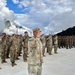 A military parade or ceremony is taking place outdoors at a mountainous location, featuring two distinct groups of soldiers in formation facing each other. Soldiers on the left wear US Army OCP (Operational Camouflage Pattern) uniforms, while soldiers on the right appear to wear a different camouflage pattern, suggesting a joint multinational military exercise or ceremony. A central figure in OCP uniform is rendering a salute, and multiple national flags are visible in the background.