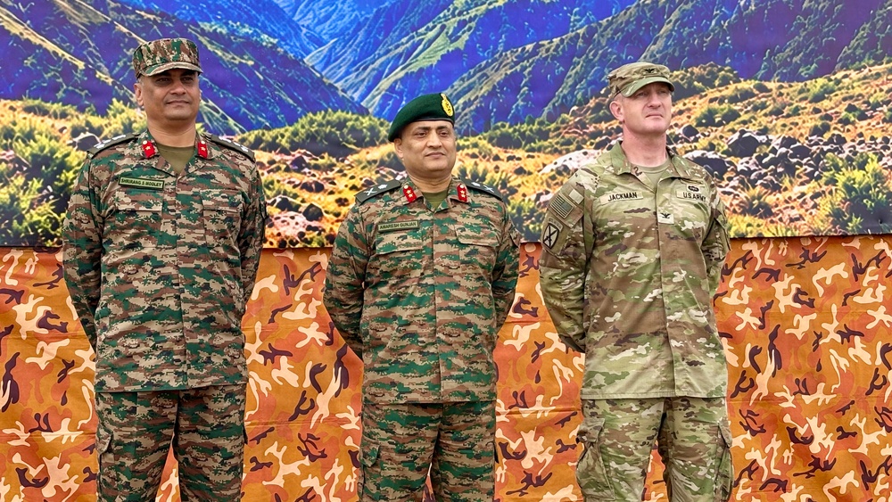 Three military officers are standing together in front of a decorative backdrop featuring a mountain landscape and camouflage pattern. Two officers are wearing Indian Army uniforms with distinctive camouflage patterns, while the third is wearing a US Army uniform. The setting appears to be a formal military event or ceremony.