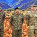Three military officers are standing together in front of a decorative backdrop featuring a mountain landscape and camouflage pattern. Two officers are wearing Indian Army uniforms with distinctive camouflage patterns, while the third is wearing a US Army uniform. The setting appears to be a formal military event or ceremony.