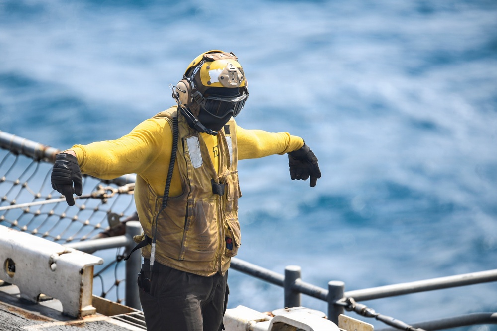 A naval deck crew member wearing yellow flight deck gear stands on what appears to be a ship's deck with the open ocean visible in the background. The individual is wearing standard carrier flight deck safety equipment including a yellow jersey, helmet with visor, black gloves, and a communications headset. Their arms are extended outward in what appears to be a hand signal gesture used for aircraft or equipment direction.