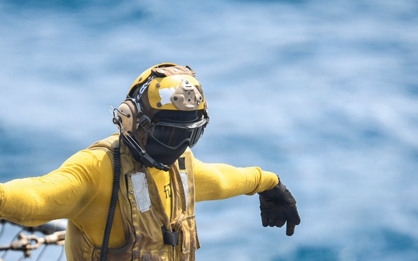 A naval deck crew member wearing yellow flight deck gear stands on what appears to be a ship's deck with the open ocean visible in the background. The individual is wearing standard carrier flight deck safety equipment including a yellow jersey, helmet with visor, black gloves, and a communications headset. Their arms are extended outward in what appears to be a hand signal gesture used for aircraft or equipment direction.