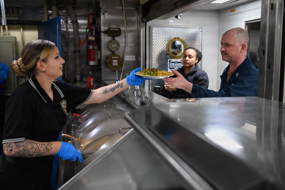 A food service worker wearing blue gloves and a black uniform hands a plate of food across a stainless steel serving counter to a male recipient in a dark blue uniform. The setting appears to be a military galley or institutional cafeteria, identifiable by the industrial equipment, fire extinguisher, and diamond-plate metal surfaces in the background. A second female worker is visible in the background observing the exchange.