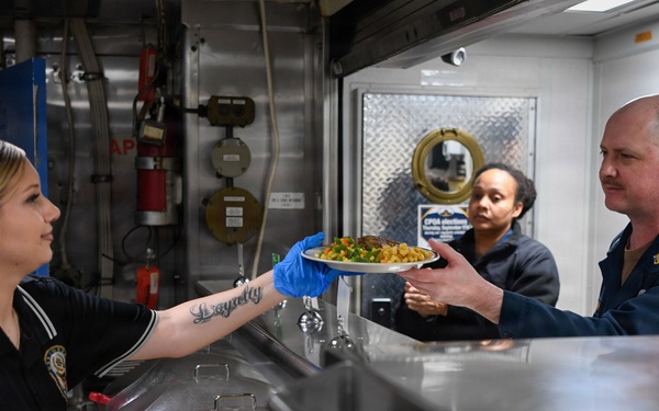 A food service worker wearing blue gloves and a black uniform hands a plate of food across a stainless steel serving counter to a male recipient in a dark blue uniform. The setting appears to be a military galley or institutional cafeteria, identifiable by the industrial equipment, fire extinguisher, and diamond-plate metal surfaces in the background. A second female worker is visible in the background observing the exchange.