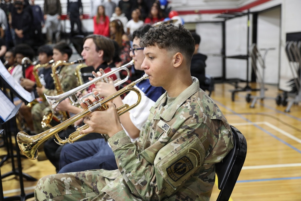 A JROTC cadet in Army OCP (camouflage) uniform is playing a trumpet in what appears to be a school gymnasium during a band performance or ceremony. Multiple musicians are seated in rows reading sheet music from stands, with a crowd of spectators visible in the bleachers behind them. The foreground cadet's uniform displays a JROTC patch on the shoulder.