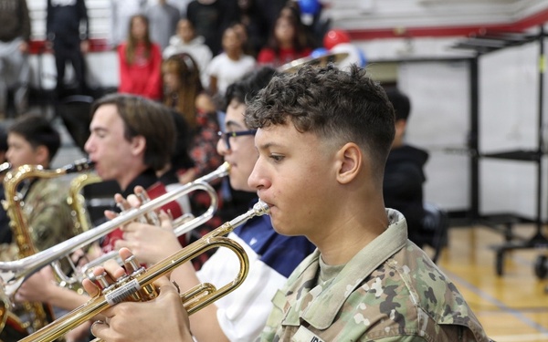 A JROTC cadet in Army OCP (camouflage) uniform is playing a trumpet in what appears to be a school gymnasium during a band performance or ceremony. Multiple musicians are seated in rows reading sheet music from stands, with a crowd of spectators visible in the bleachers behind them. The foreground cadet's uniform displays a JROTC patch on the shoulder.