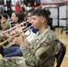 A JROTC cadet in Army OCP (camouflage) uniform is playing a trumpet in what appears to be a school gymnasium during a band performance or ceremony. Multiple musicians are seated in rows reading sheet music from stands, with a crowd of spectators visible in the bleachers behind them. The foreground cadet's uniform displays a JROTC patch on the shoulder.