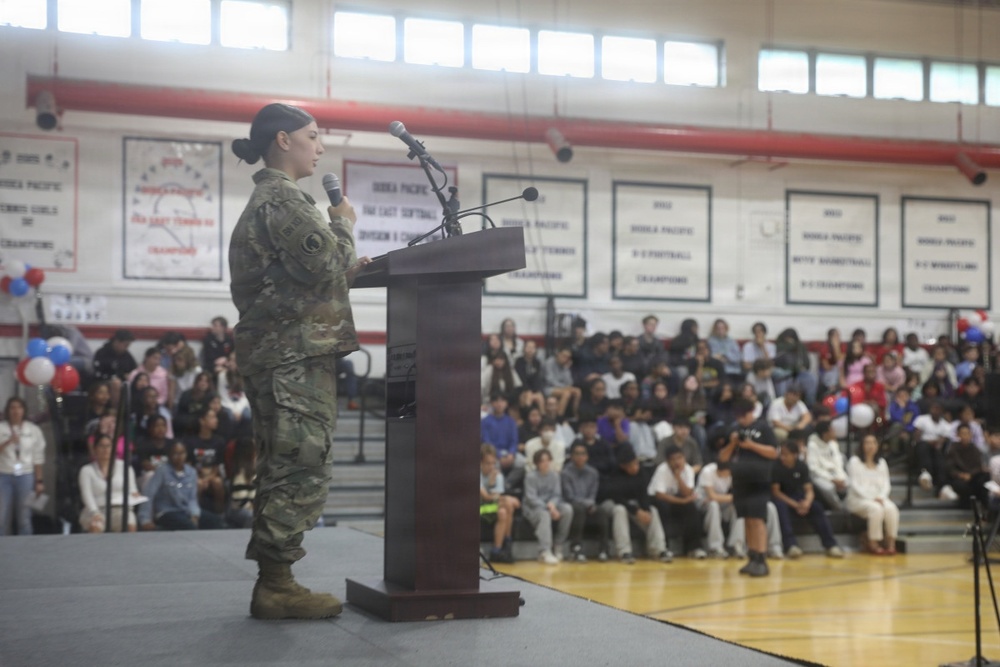 A female soldier in U.S. Army camouflage (OCP) uniform is speaking at a podium on a gymnasium stage to a large seated audience. The setting appears to be a school gymnasium, likely during a Veterans Day or military appreciation assembly. Championship banners referencing 'Korea Pacific' competitions are visible on the walls, suggesting this may be a Department of Defense Dependents School (DoDDS).