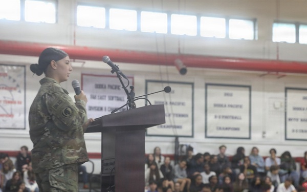 A female soldier in U.S. Army camouflage (OCP) uniform is speaking at a podium on a gymnasium stage to a large seated audience. The setting appears to be a school gymnasium, likely during a Veterans Day or military appreciation assembly. Championship banners referencing 'Korea Pacific' competitions are visible on the walls, suggesting this may be a Department of Defense Dependents School (DoDDS).