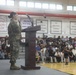 A female soldier in U.S. Army camouflage (OCP) uniform is speaking at a podium on a gymnasium stage to a large seated audience. The setting appears to be a school gymnasium, likely during a Veterans Day or military appreciation assembly. Championship banners referencing 'Korea Pacific' competitions are visible on the walls, suggesting this may be a Department of Defense Dependents School (DoDDS).