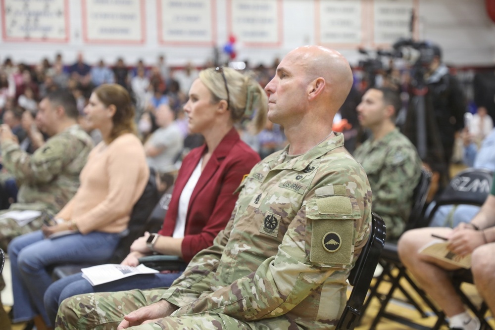 A group of U.S. Army soldiers and civilians are seated in what appears to be a gymnasium or indoor arena during a formal military ceremony or event. The foreground features a bald male soldier in OCP (Operational Camouflage Pattern) uniform with visible U.S. Army tape and unit patches. The audience includes both uniformed personnel and civilian attendees, with media equipment visible in the background.
