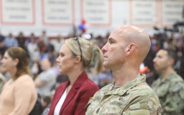 A group of U.S. Army soldiers and civilians are seated in what appears to be a gymnasium or indoor arena during a formal military ceremony or event. The foreground features a bald male soldier in OCP (Operational Camouflage Pattern) uniform with visible U.S. Army tape and unit patches. The audience includes both uniformed personnel and civilian attendees, with media equipment visible in the background.