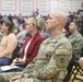 A group of U.S. Army soldiers and civilians are seated in what appears to be a gymnasium or indoor arena during a formal military ceremony or event. The foreground features a bald male soldier in OCP (Operational Camouflage Pattern) uniform with visible U.S. Army tape and unit patches. The audience includes both uniformed personnel and civilian attendees, with media equipment visible in the background.