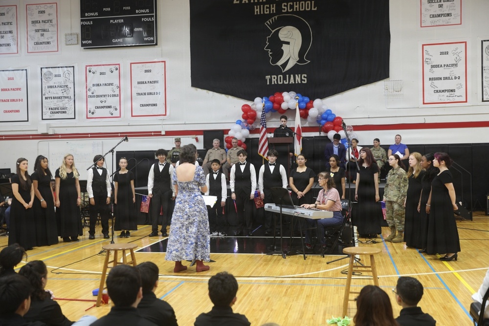 A school choir performance is taking place in a high school gymnasium identified as a 'Trojans' school, likely a patriotic ceremony such as Veterans Day or a similar military appreciation event. Students dressed in black formal attire are performing while a conductor in a floral dress leads them, accompanied by a keyboardist. Military personnel in uniform are visible in the background near an American flag and red, white, and blue balloon arch.