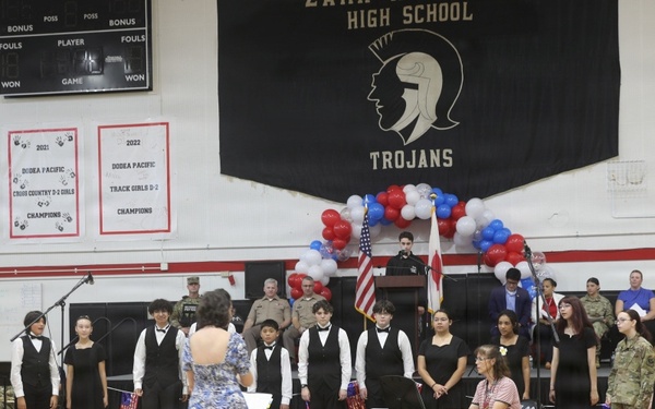 A school choir performance is taking place in a high school gymnasium identified as a 'Trojans' school, likely a patriotic ceremony such as Veterans Day or a similar military appreciation event. Students dressed in black formal attire are performing while a conductor in a floral dress leads them, accompanied by a keyboardist. Military personnel in uniform are visible in the background near an American flag and red, white, and blue balloon arch.