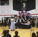 A school choir performance is taking place in a high school gymnasium identified as a 'Trojans' school, likely a patriotic ceremony such as Veterans Day or a similar military appreciation event. Students dressed in black formal attire are performing while a conductor in a floral dress leads them, accompanied by a keyboardist. Military personnel in uniform are visible in the background near an American flag and red, white, and blue balloon arch.