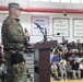 A U.S. Army soldier in full camouflage uniform is speaking at a podium equipped with microphones in what appears to be a school gymnasium. The audience consists of a large group of students seated in bleachers, suggesting this is a school assembly, likely related to Veterans Day or a military appreciation event. Red, white, and blue balloons are visible, reinforcing a patriotic theme.