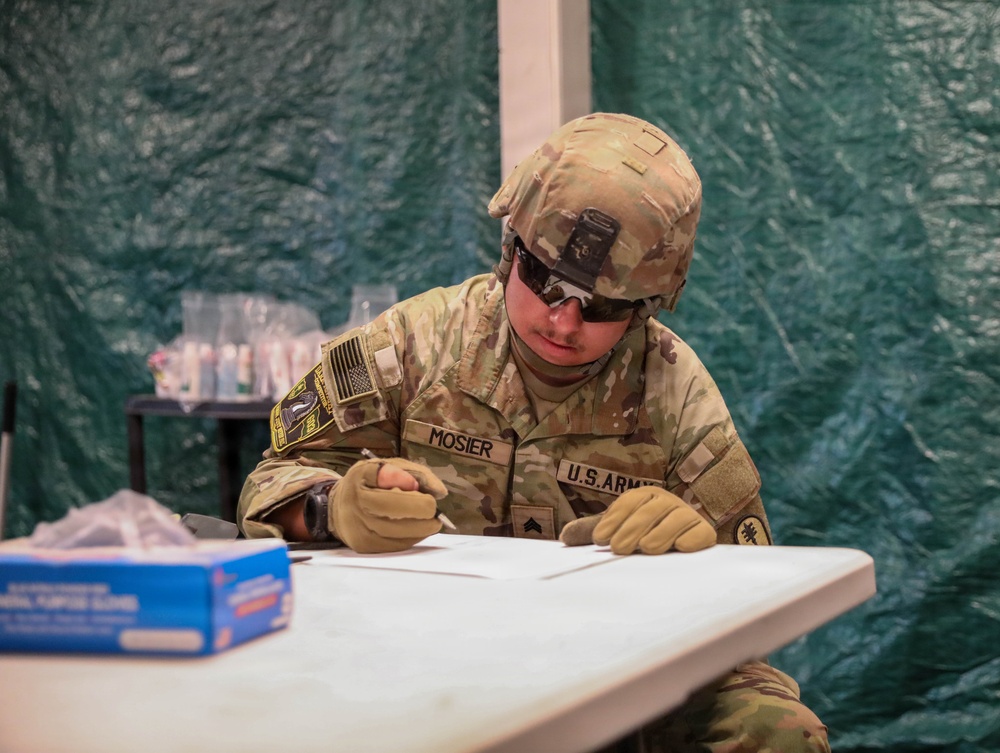 A U.S. Army soldier named Mosier, wearing Operational Camouflage Pattern (OCP) uniform, combat helmet with NVG mount, tactical gloves, and sunglasses, is seated at a white folding table writing on documents. The setting appears to be inside a military field tent or temporary structure with green tarpaulin walls. A blue box of bandages and other medical or administrative supplies are visible on the table nearby.