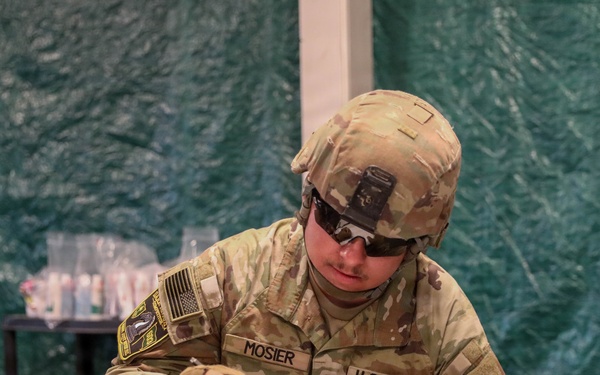 A U.S. Army soldier named Mosier, wearing Operational Camouflage Pattern (OCP) uniform, combat helmet with NVG mount, tactical gloves, and sunglasses, is seated at a white folding table writing on documents. The setting appears to be inside a military field tent or temporary structure with green tarpaulin walls. A blue box of bandages and other medical or administrative supplies are visible on the table nearby.