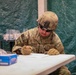 A U.S. Army soldier named Mosier, wearing Operational Camouflage Pattern (OCP) uniform, combat helmet with NVG mount, tactical gloves, and sunglasses, is seated at a white folding table writing on documents. The setting appears to be inside a military field tent or temporary structure with green tarpaulin walls. A blue box of bandages and other medical or administrative supplies are visible on the table nearby.