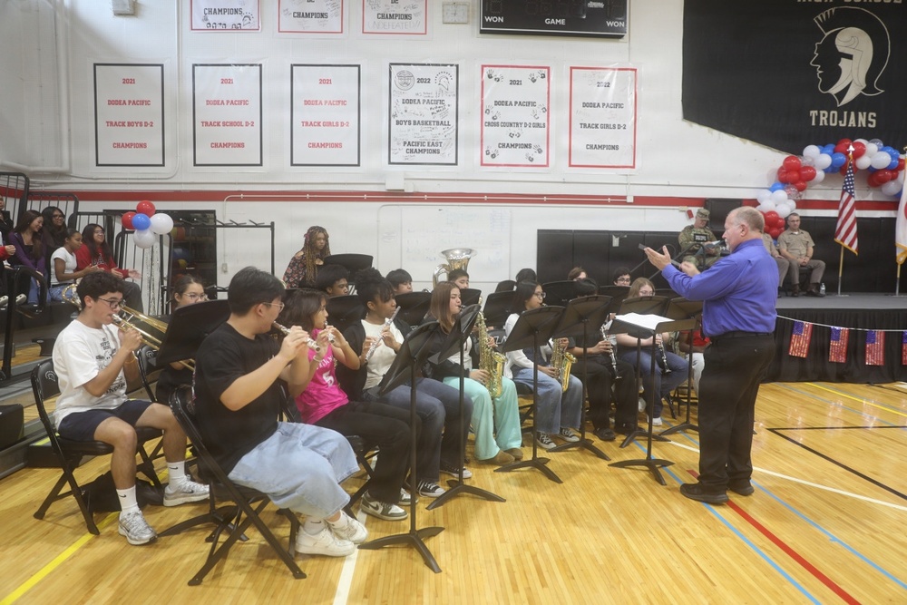 A school band performs in a gymnasium under the direction of a conductor wearing a blue shirt. The students are seated in a semicircular arrangement playing various instruments including trumpets, flutes, saxophones, and clarinets. The setting appears to be a school assembly or ceremony, likely patriotic in nature given the American flag and red, white, and blue decorations visible.