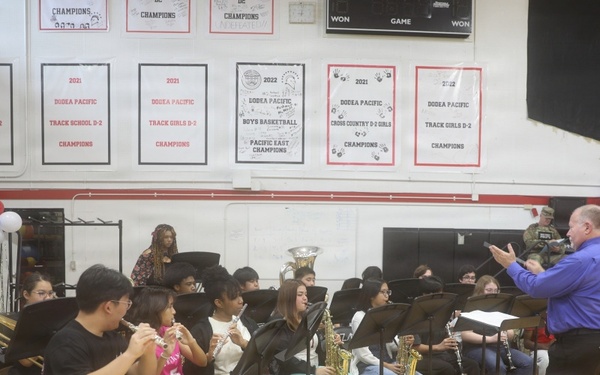 A school band performs in a gymnasium under the direction of a conductor wearing a blue shirt. The students are seated in a semicircular arrangement playing various instruments including trumpets, flutes, saxophones, and clarinets. The setting appears to be a school assembly or ceremony, likely patriotic in nature given the American flag and red, white, and blue decorations visible.