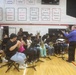 A school band performs in a gymnasium under the direction of a conductor wearing a blue shirt. The students are seated in a semicircular arrangement playing various instruments including trumpets, flutes, saxophones, and clarinets. The setting appears to be a school assembly or ceremony, likely patriotic in nature given the American flag and red, white, and blue decorations visible.