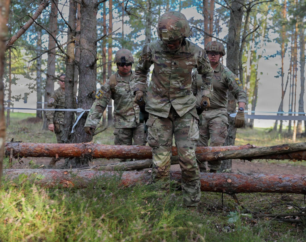 A group of U.S. Army soldiers in full combat gear and MultiCam (OCP) uniforms are navigating a woodland obstacle course, stepping over felled logs. The lead soldier has a visible name tape reading 'U.S. ARMY' and appears to be carefully crossing a log barrier. The setting appears to be a military training area in a pine forest, possibly in Europe based on the terrain and vegetation.
