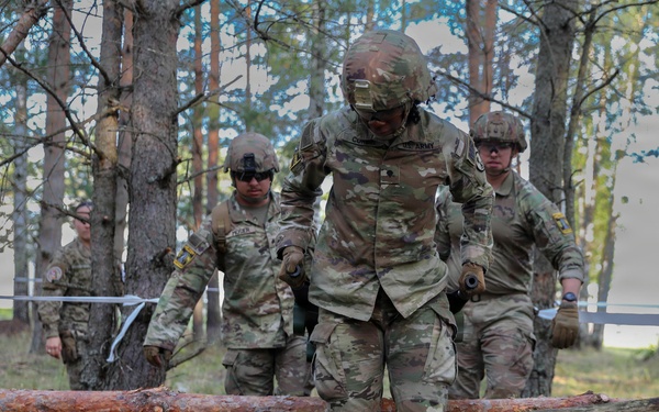 A group of U.S. Army soldiers in full combat gear and MultiCam (OCP) uniforms are navigating a woodland obstacle course, stepping over felled logs. The lead soldier has a visible name tape reading 'U.S. ARMY' and appears to be carefully crossing a log barrier. The setting appears to be a military training area in a pine forest, possibly in Europe based on the terrain and vegetation.