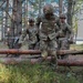 A group of U.S. Army soldiers in full combat gear and MultiCam (OCP) uniforms are navigating a woodland obstacle course, stepping over felled logs. The lead soldier has a visible name tape reading 'U.S. ARMY' and appears to be carefully crossing a log barrier. The setting appears to be a military training area in a pine forest, possibly in Europe based on the terrain and vegetation.