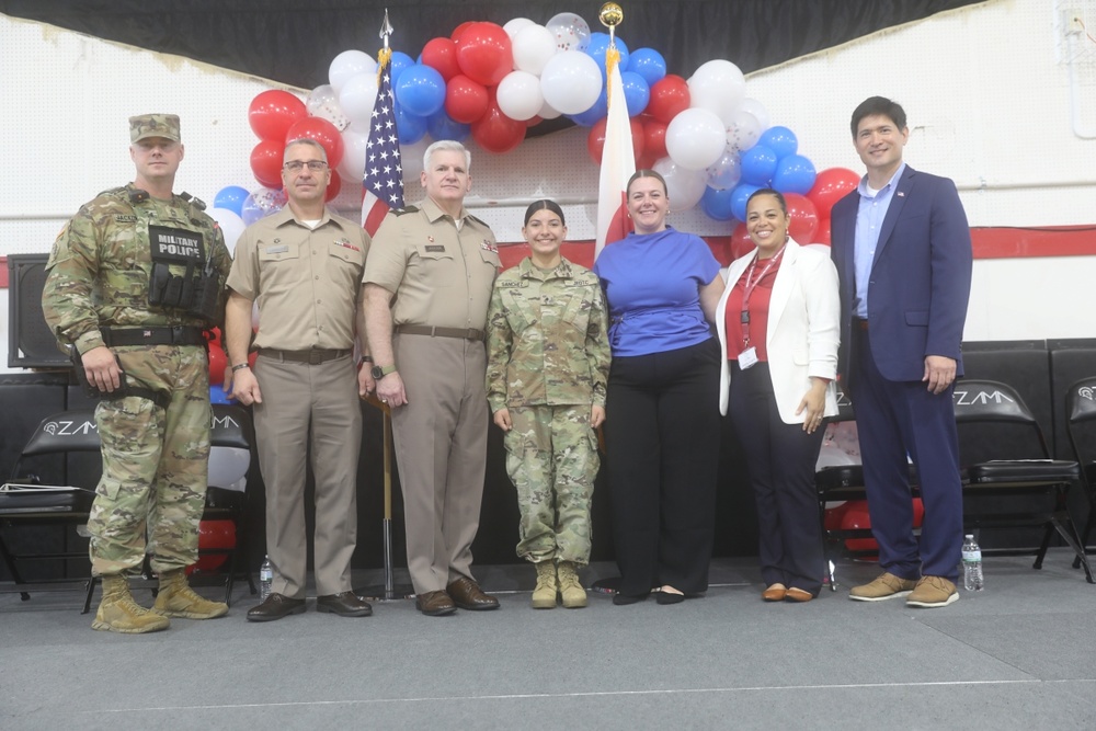 A group of seven individuals pose for a formal photo in front of a patriotic red, white, and blue balloon arch with an American flag. The group includes four military personnel in uniform and three civilians, suggesting an official military ceremony or event. The setting appears to be an indoor venue decorated for a patriotic occasion.
